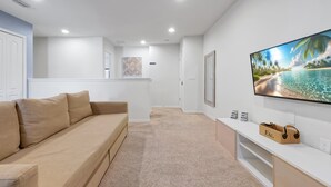 A living room with a beige sofa, a flat-screen TV mounted on the wall, a white entertainment unit, and a carpeted floor.