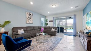 A modern living room with a sectional sofa, a coffee table, and a blue armchair. There is a glass door leading to a balcony with a view of a pool.
