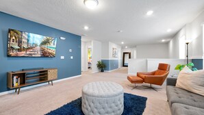A modern living room with a blue accent wall, a large flat-screen TV, a leather armchair, a grey ottoman, and a bookshelf. - 7BR Solterra Villa | Private Pool & Spa | Mickey Room | Near Disney (Davenport)