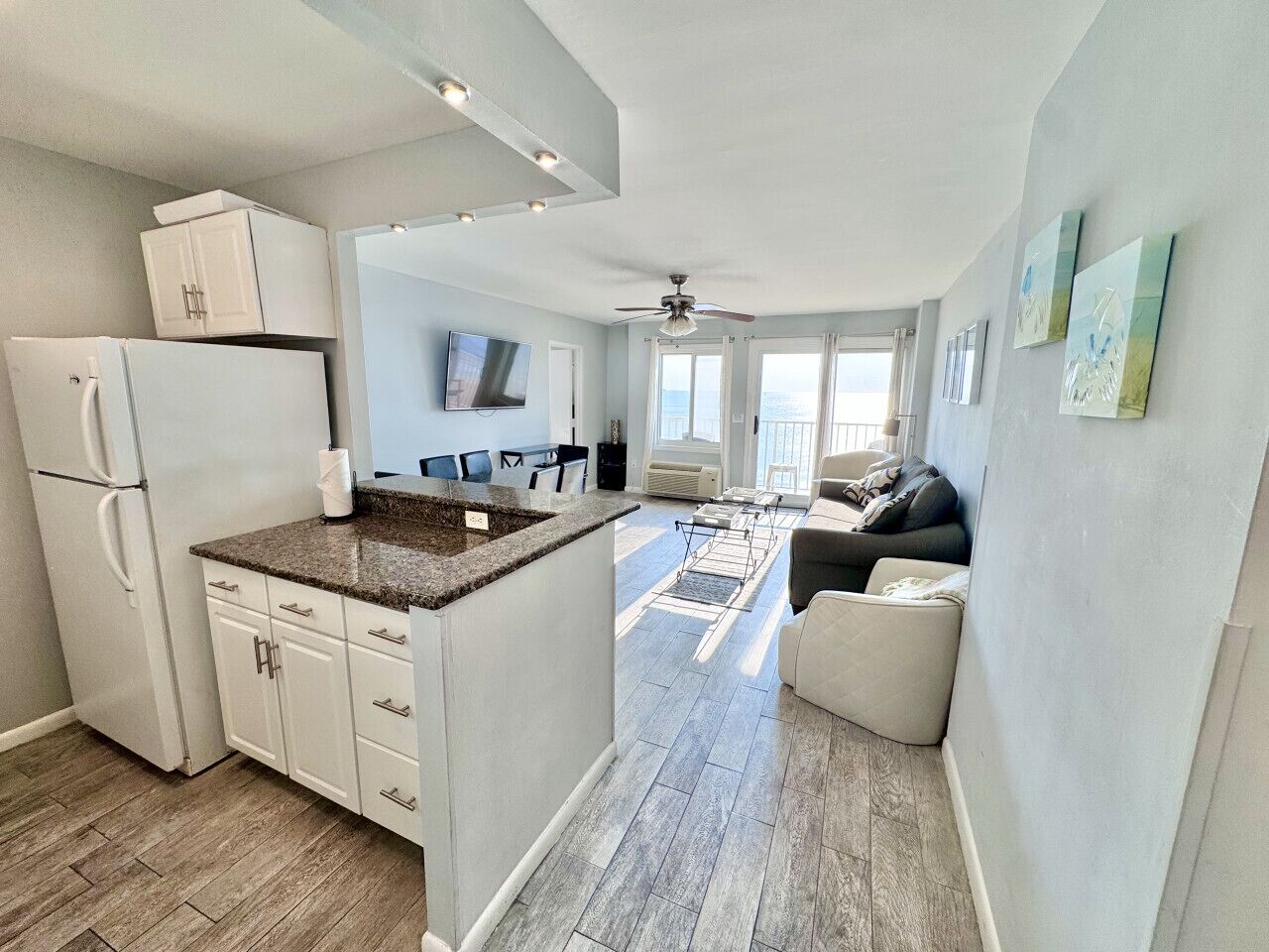 A modern kitchen with a built-in island, white cabinetry, and a refrigerator. There is a dining area with a sofa and a view of the outdoors.