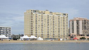 A high-rise hotel with multiple balconies, a beach in the foreground, and several white tents set up on the sand. - Beach House Oceanfront Condo | Indoor Pool Heated 606 (Virginia Beach)
