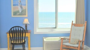 A room with a large window overlooking the sea, a chair, a table with a lamp, and a framed picture on the wall. - Beach House Oceanfront Condo | Indoor Pool Heated 606 (Virginia Beach)