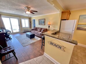 A kitchen with a countertop labeled "BEACH," a dining area with chairs, a sofa, and a ceiling fan. - Beach House Oceanfront Condo | Indoor Pool Heated (Virginia Beach)