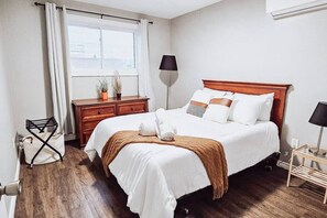 A neatly made bed with white bedding and a brown throw, a wooden nightstand with a lamp, a window with curtains, and a small table with a lamp. - Spacious 3BR Retreat, Firepit & Outdoor Kitchen – Walk to Falls (Niagara Falls)
