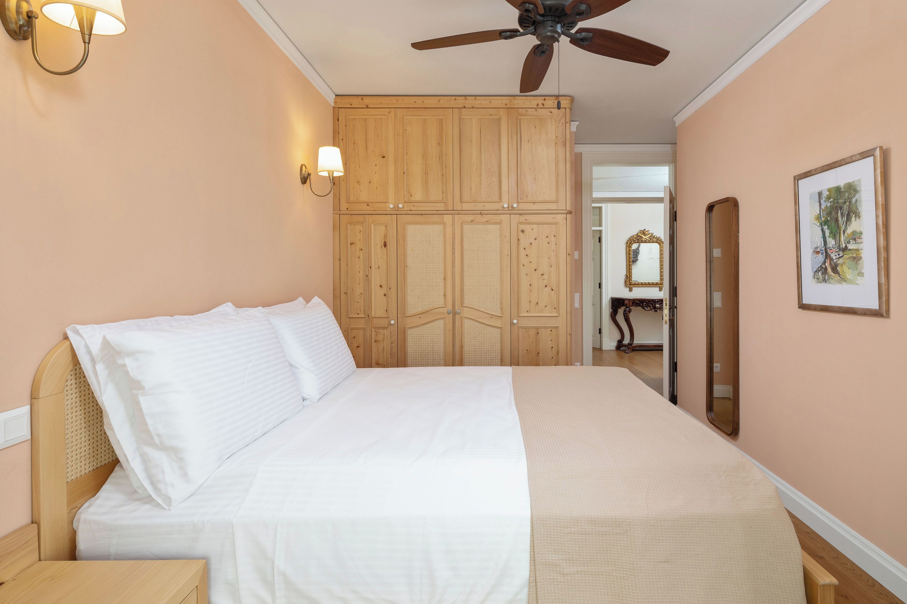 A bedroom with a bed, wooden wardrobe, ceiling fan, wall-mounted lamp, and a framed picture.