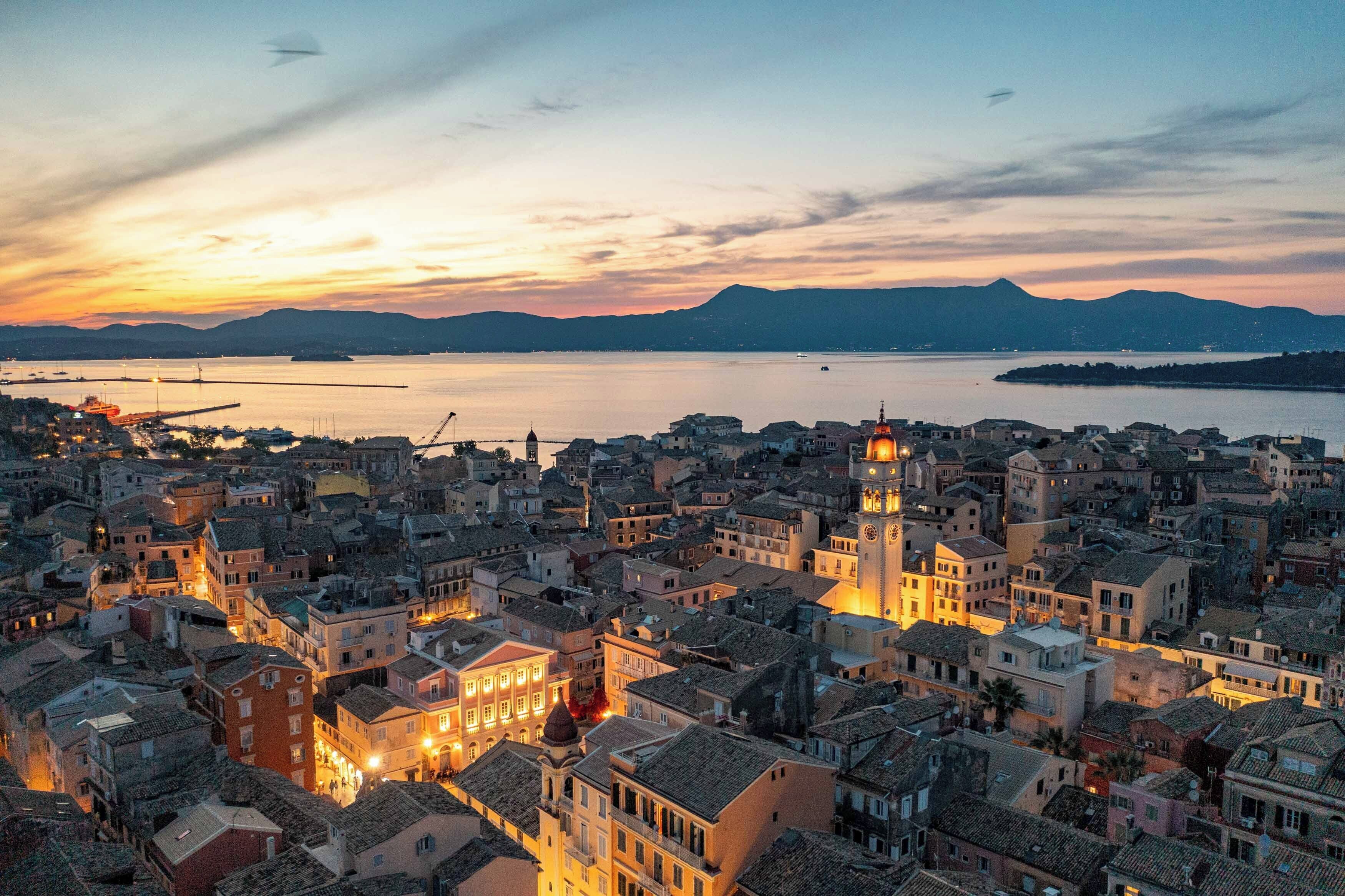 A coastal cityscape at sunset with historic buildings, a prominent clock tower, and a calm sea.