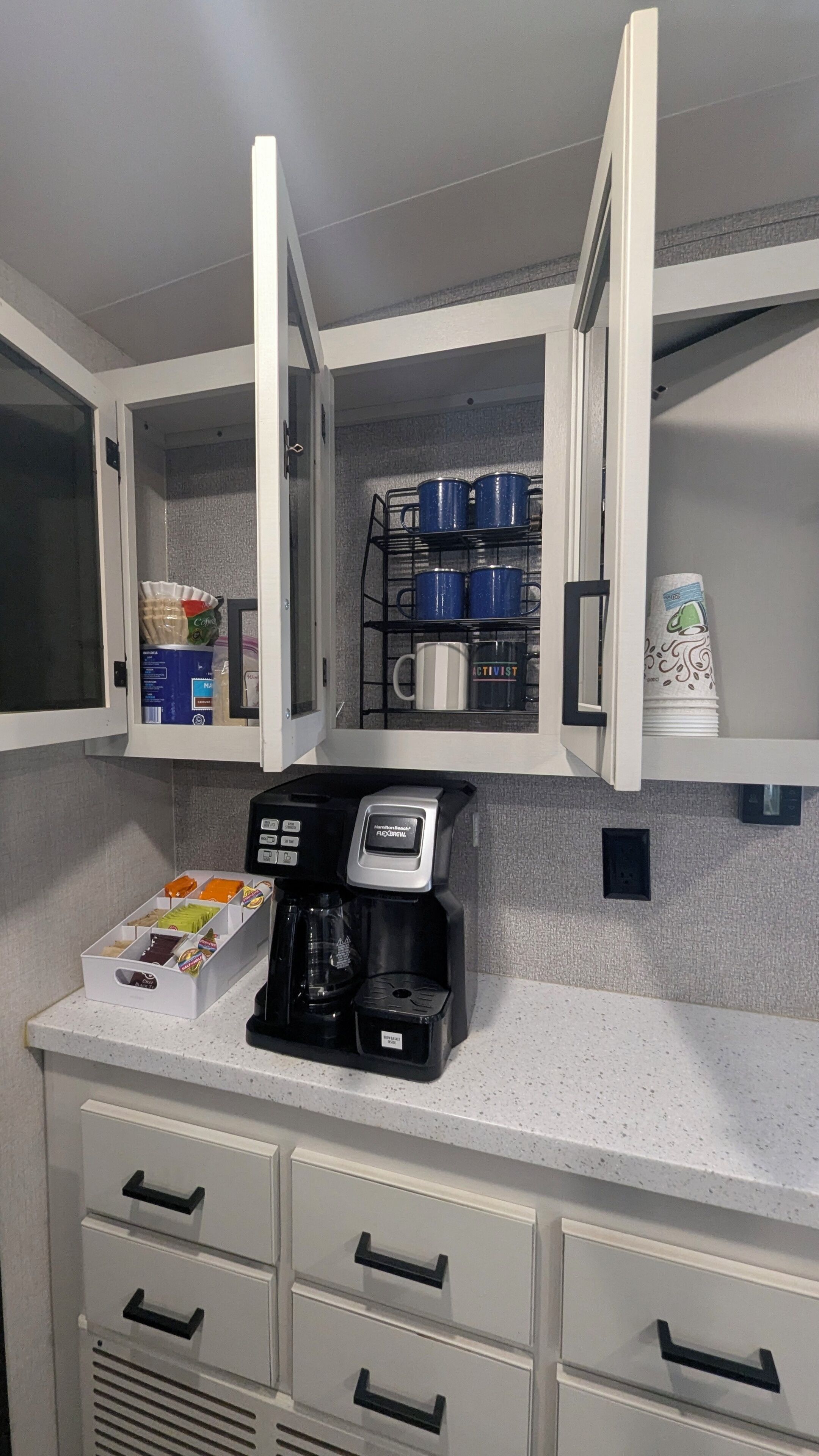 A kitchenette with a coffee maker, storage cabinets, and a shelf with blue mugs.