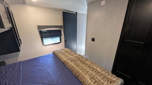 Une petite pièce comprenant un lit, une fenêtre avec des stores, une porte et une petite fenêtre située au-dessus du lit.