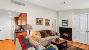A living room with a sofa, a coffee table, and a fireplace. There is a kitchen area with cabinets and a stove. - Elevate your Escape (Lake George)