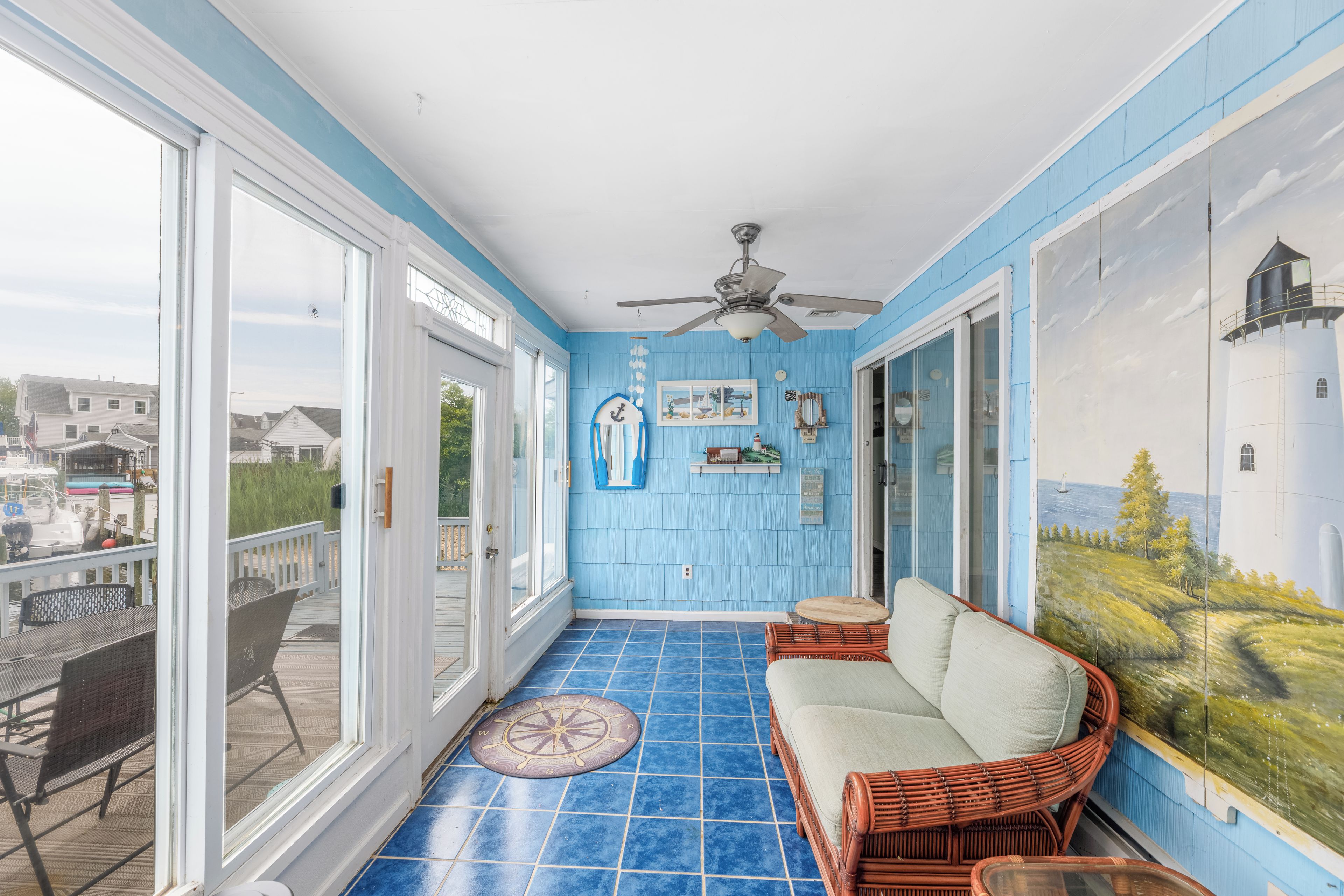 A sunlit room with a blue tiled floor, a ceiling fan, a wicker sofa, and a large window overlooking a dock and boats.