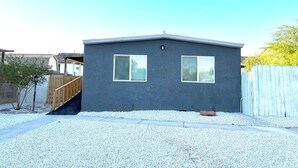 Una casa pequeña de un solo piso con fachada gris oscuro, dos ventanas con contraventanas blancas y una cerca de madera al fondo.