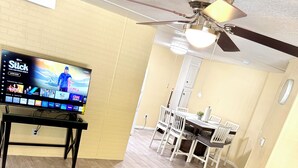 Una sala de estar con televisor sobre un mueble bajo, ventilador de techo, comedor con mesa y sillas, y una alfombra mullida.