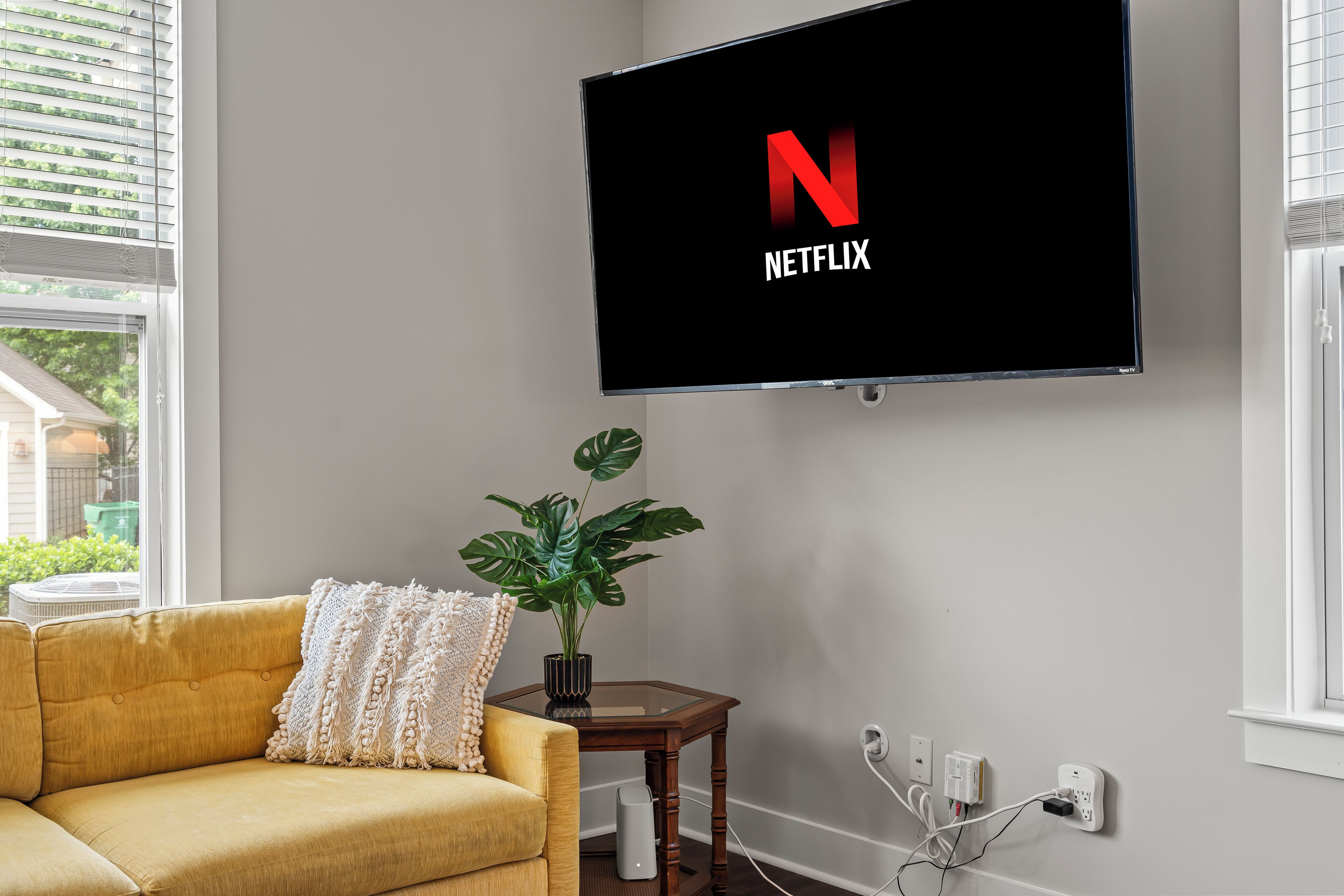 A modern living room with a mounted TV displaying the Netflix logo, a yellow sofa, a potted plant, and a small wooden side table.