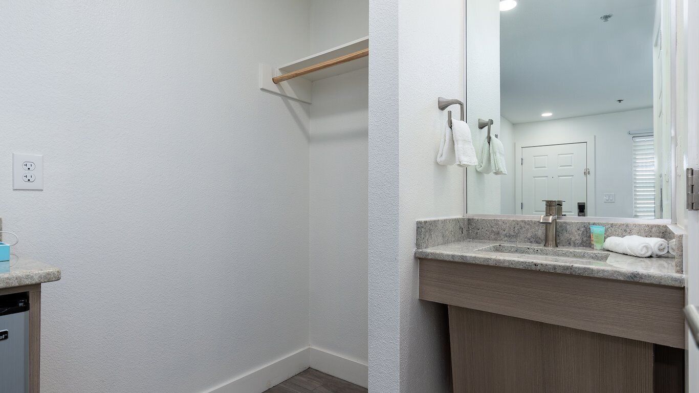A bathroom with a white wall, a wooden shelf, a sink with a mirror, and a door in the background.