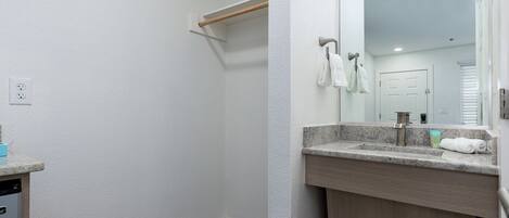 A bathroom with a white wall, a wooden shelf, a sink with a mirror, and a door in the background.