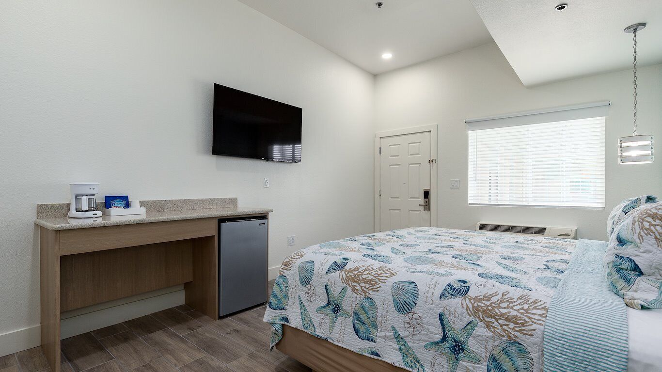 A bedroom with a bed, a TV mounted on the wall, a small desk with a coffee maker, and a window with blinds.