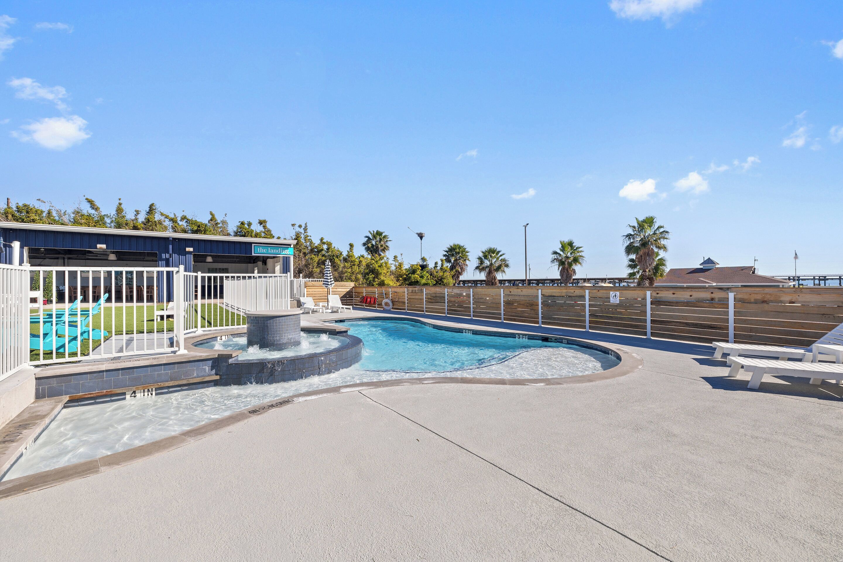 A swimming pool with a water feature, surrounded by a white fence and palm trees.