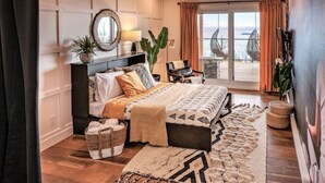 Une chambre avec un grand lit, une table de chevet, un miroir, une plante en pot, une chaise et une vue sur la mer, visible à travers une porte-fenêtre coulissante.