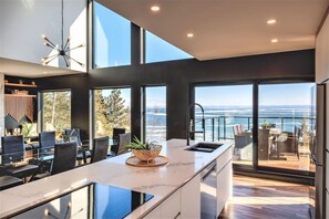 Une cuisine moderne dotée d’un grand îlot central, d’un coin repas avec des chaises et d’une vue sur l’océan grâce à de grandes fenêtres.
