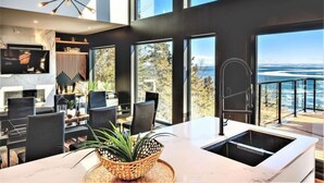 Une cuisine moderne dotée d’un grand îlot central, d’un coin repas avec des chaises noires et de grandes fenêtres offrant une vue sur l’océan.