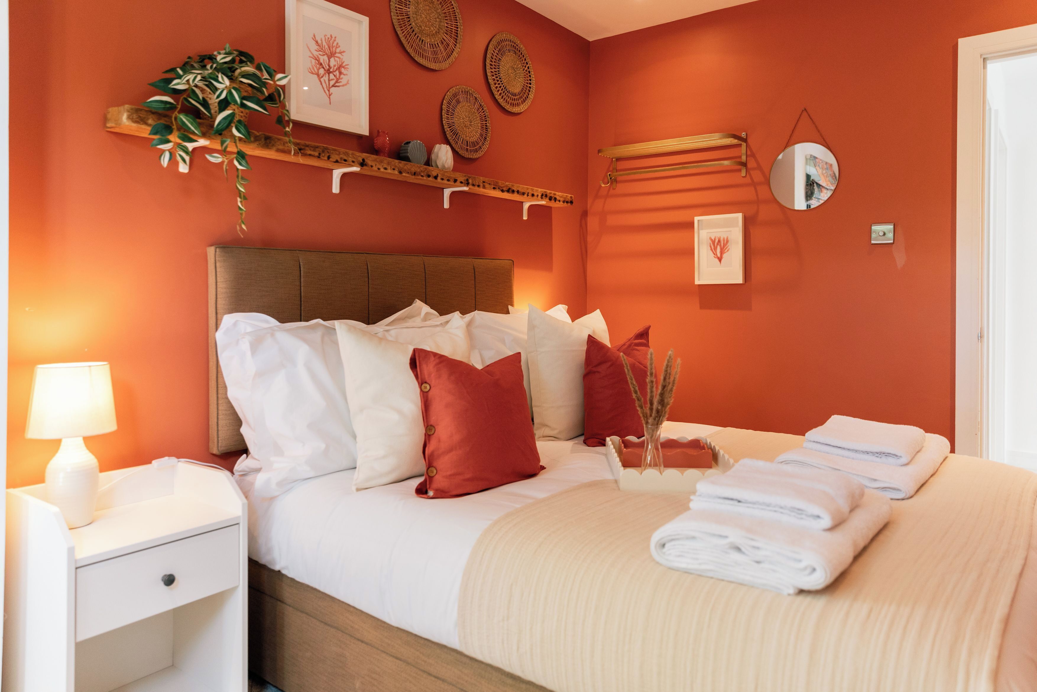 A neatly made bed with white and red pillows, a bedside lamp, and a small plant on a nightstand.