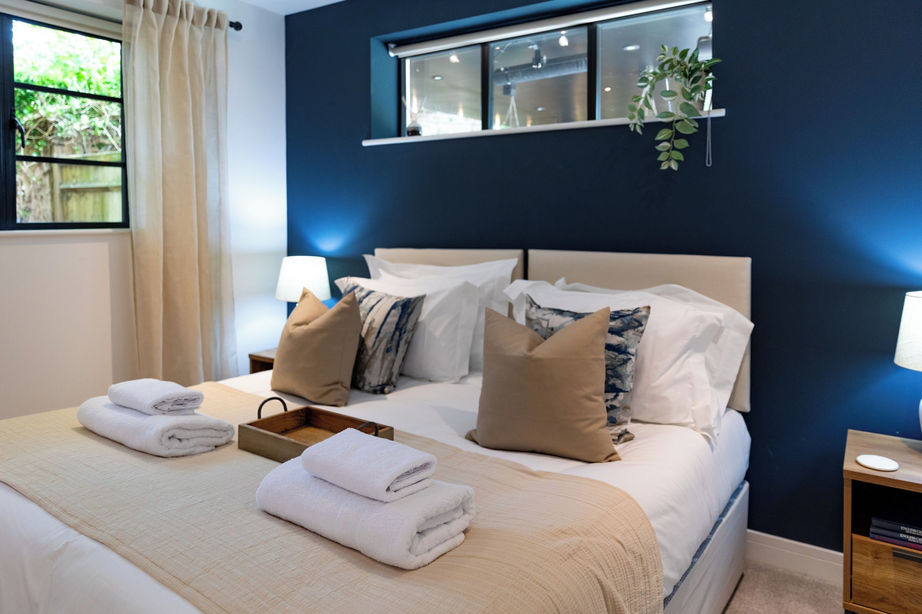 A neatly made bed with white linens and a beige throw blanket, a bedside table with a lamp, and a window with curtains.