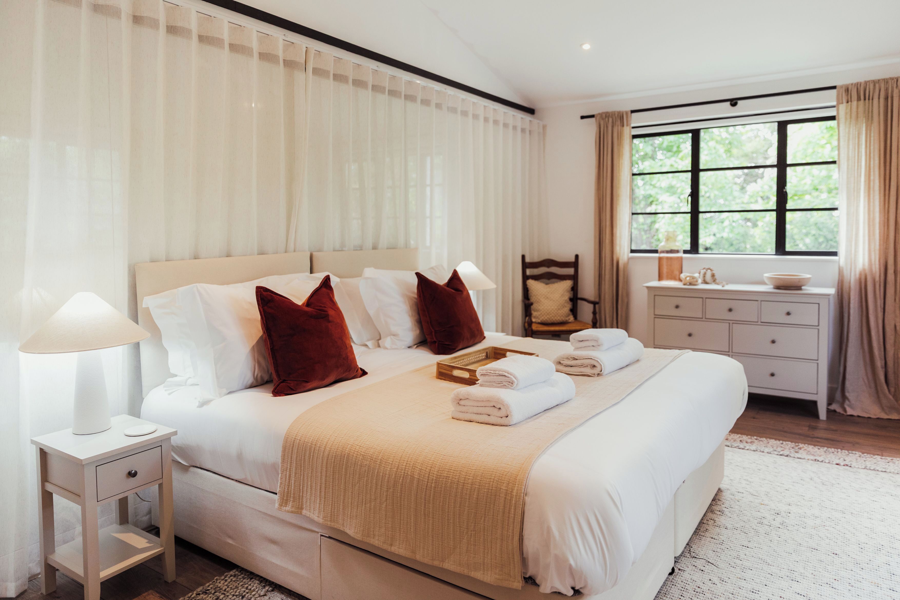 Una cama bien tendida con sábanas blancas y dos almohadas rojas, una mesita de noche con una lámpara, una silla de madera, una pequeña mesa con un cuenco y una ventana con cortinas.