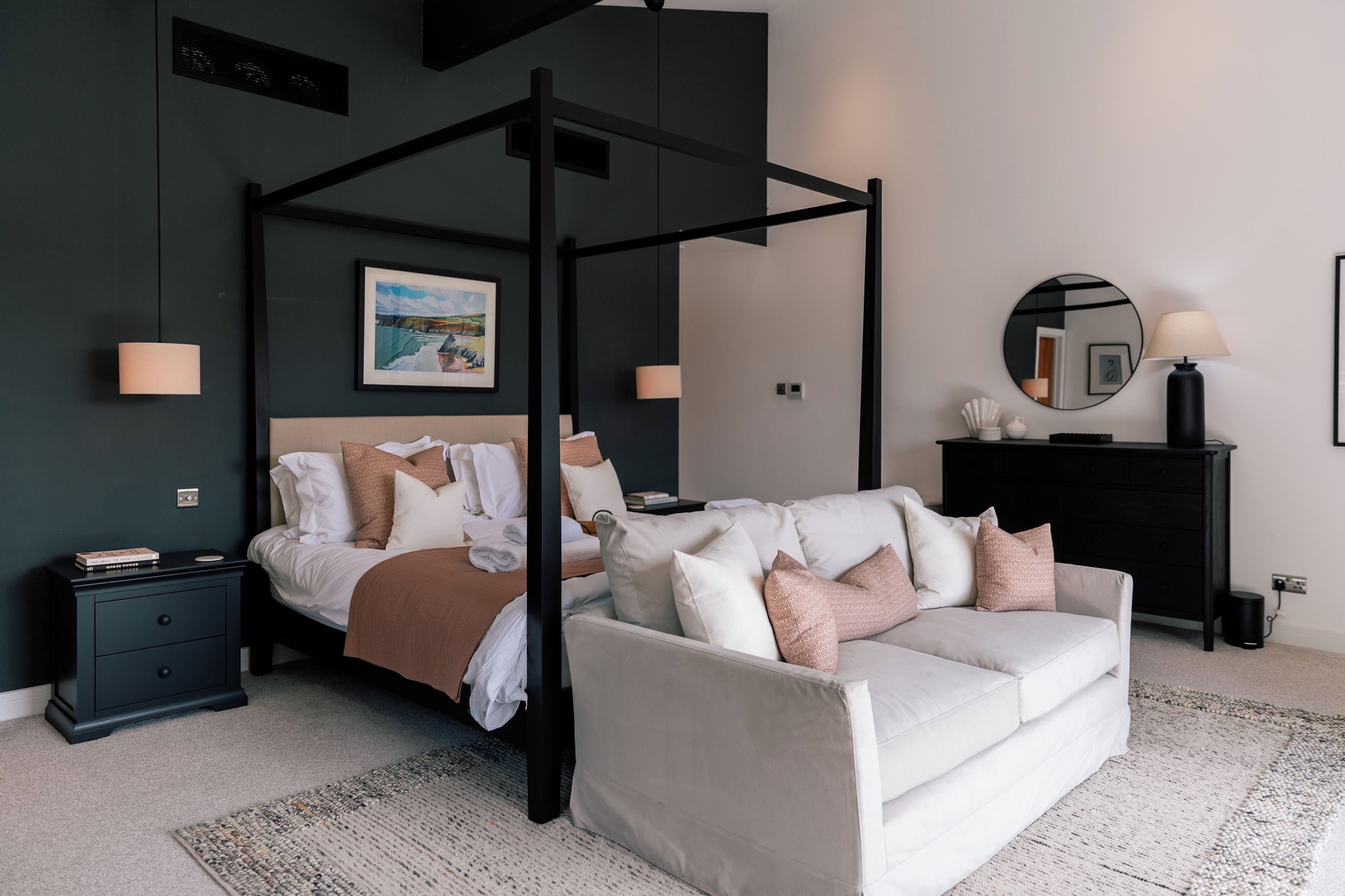 Un dormitorio con cama con dosel, un sofá blanco, una mesita de noche negra, un aparador negro, un espejo redondo y un cuadro en la pared.