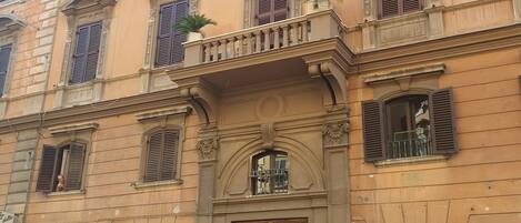 A multi-story building with a central entrance, potted plants on the balcony, and closed shutters on the windows.