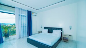 Quarto de hotel moderno com uma cama grande, uma mesa de cabeceira, ar condicionado e vista para a cidade através de uma janela ampla.