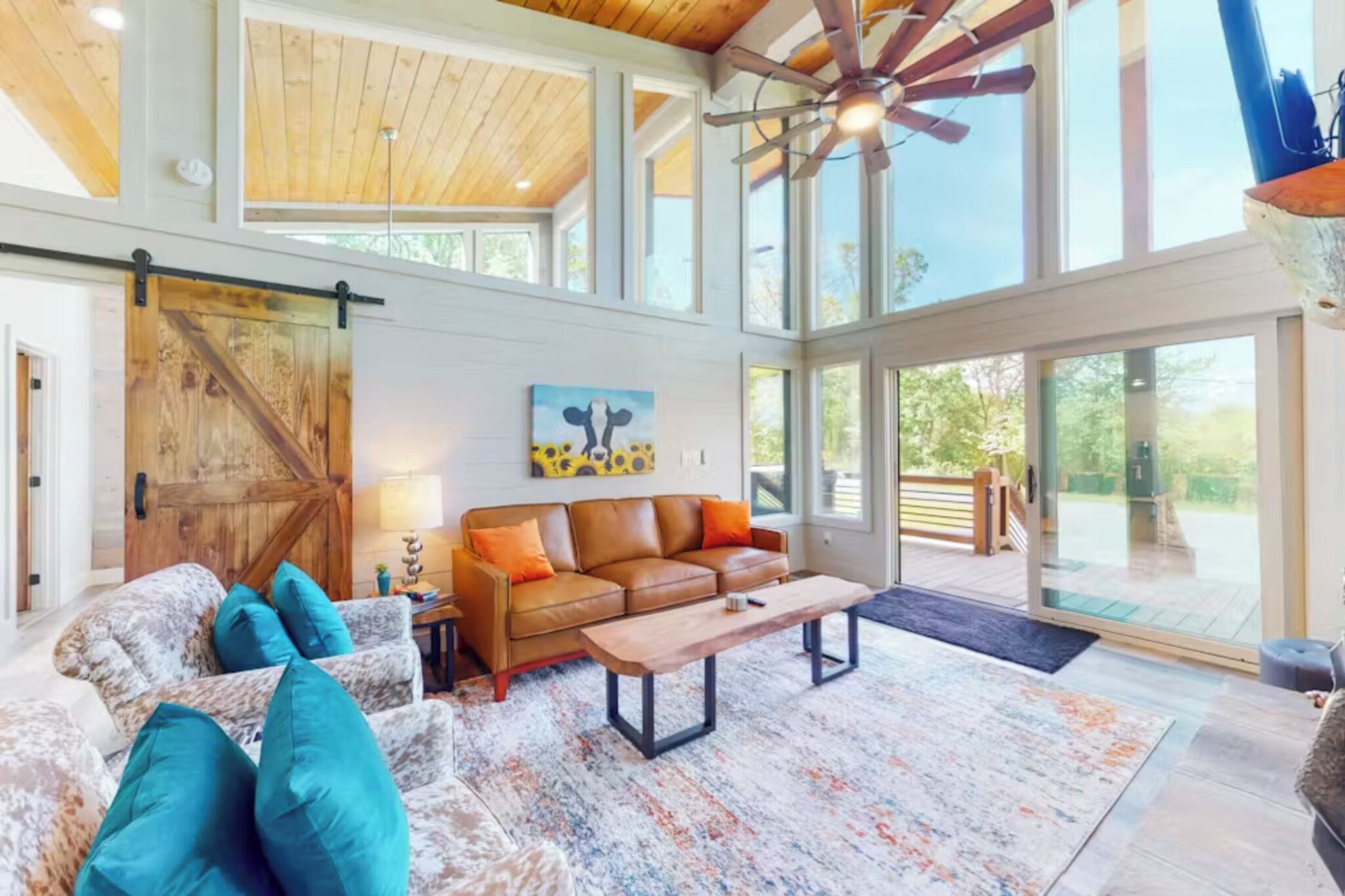 A spacious living room with a large sliding barn door, a brown leather sofa, teal cushions, a wooden coffee table, and a ceiling fan.
