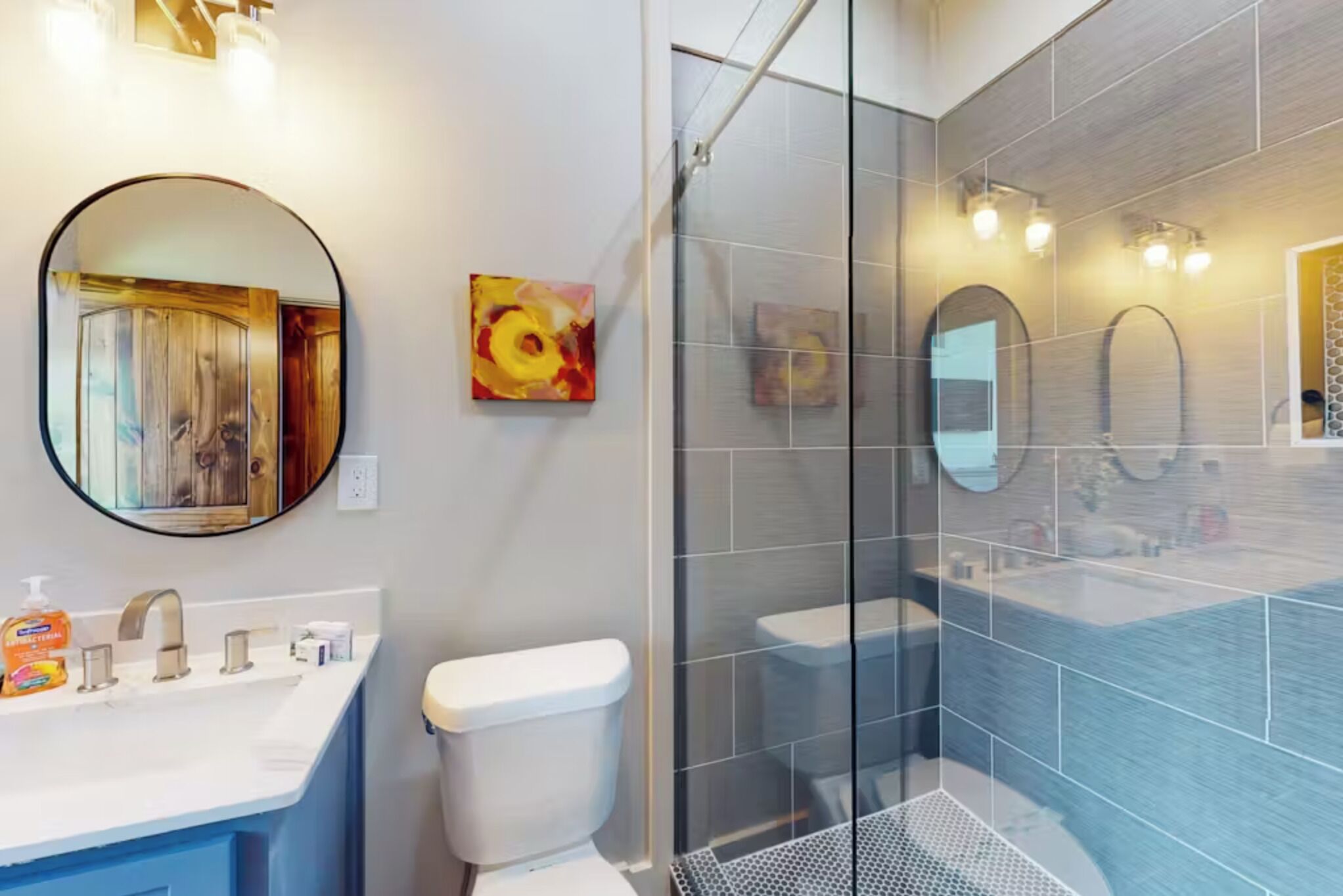 A modern bathroom with a glass-enclosed shower, a round mirror, and a white sink with a blue cabinet.