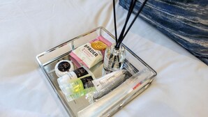 A bedside table with a glass tray containing toiletries, a bottle of perfume, and a pack of tissues.