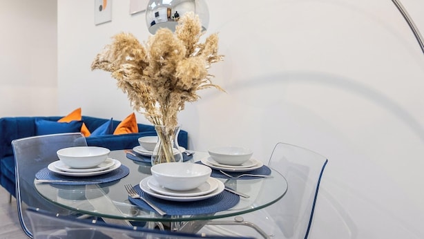 A modern dining area with a glass table set for four, surrounded by blue sofas and a decorative vase with dried plants.