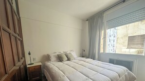 A bedroom with a large bed, a nightstand, a lamp, and a window with curtains. - Rent Up -Monoambiente en zona tribunales (Buenos Aires)