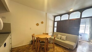 A room with a wooden herringbone floor, a round wooden table with chairs, a beige sofa, and a large window with a view. - Rent Up -Monoambiente en zona tribunales (Buenos Aires)