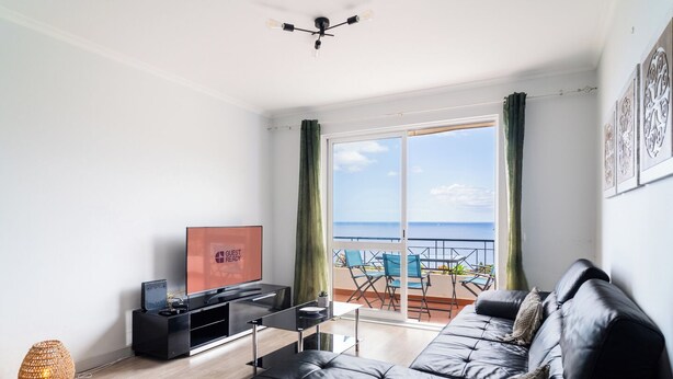 A modern living room with a black leather sofa, a flat-screen TV, and a balcony with a view of the sea.