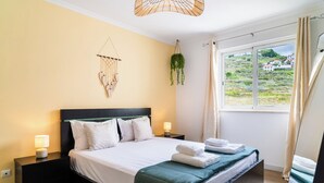 A neatly made bed with white linens and a green throw, a bedside table with a lamp, a macrame wall hanging, and a view of a green landscape through a window.