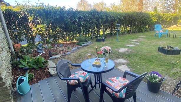 A deck with a table and chairs, surrounded by a garden with a hedge and potted plants. - Chez Sandrine (La Gacilly)
