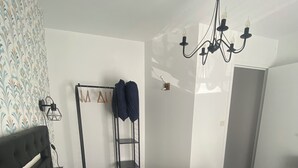 A bedroom with a patterned bedspread, a black headboard, a wall-mounted light fixture, and a clothes rack with hanging garments. - Chez Sandrine (La Gacilly)