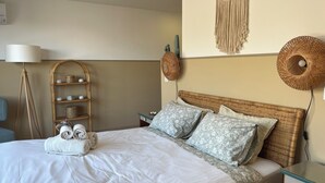 Una cama bien tendida con toallas enrolladas, una mesita de noche de mimbre, una lámpara y decoraciones de macramé sujetas a la pared.