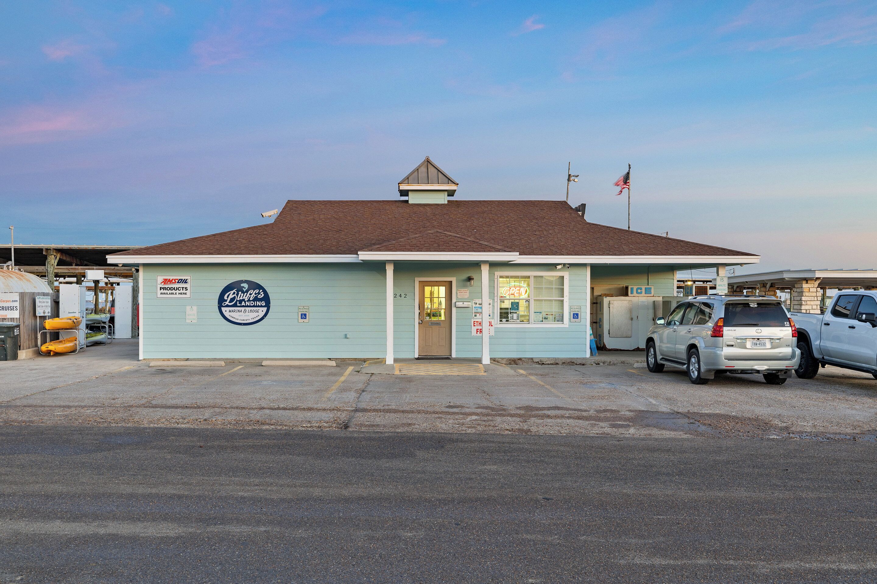A small, light blue building with a brown roof, a sign reading "Buch's Landing," and a flag on top. There are two cars parked in front.