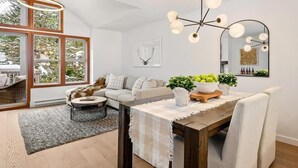 A modern living room with a dining area, featuring a wooden dining table, a sofa, and a large window with a view of the outdoors. - Updated Ski In 3-bedroom townhome in Whistler (Whistler)