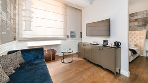 A modern living room with a dark blue sofa, a small round table, and a grey cabinet. A large window with blinds allows natural light to enter. - Rhea Silvia Luxury Rooms Spagna (Rome)