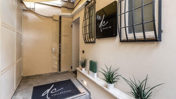 A hotel entrance with a doormat, a reception desk, a window with a metal grill, and potted plants on a shelf. - Rhea Silvia Luxury Rooms Spagna (Rome)