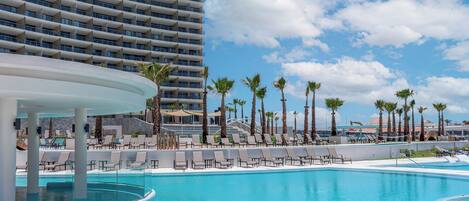 A resort pool area with lounge chairs and palm trees, surrounded by a high-rise building.