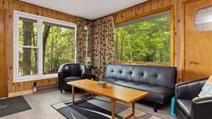 Une pièce chaleureuse avec un décor intérieur en bois, une grande fenêtre donnant sur la forêt, un canapé en cuir noir et une table basse en bois.