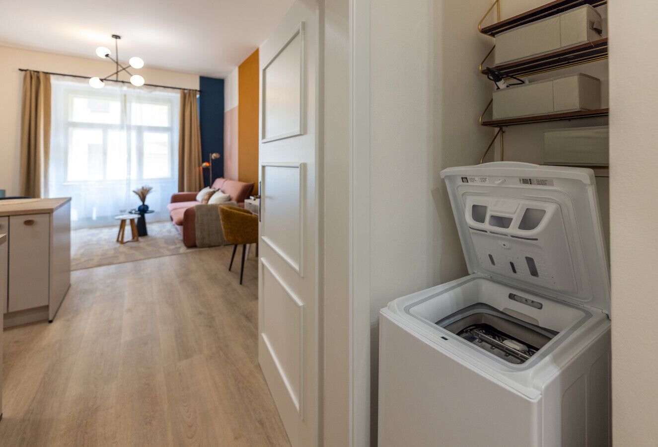 A laundry room with a front-loading washing machine, a wooden floor, a dining area with a table and chairs, and a window with curtains.