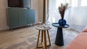 Un salon moderne avec un téléviseur à écran plat posé sur un meuble, une table ronde en bois avec deux verres à vin, un vase bleu contenant des plantes séchées et une grande fenêtre ornée de rideaux légers.