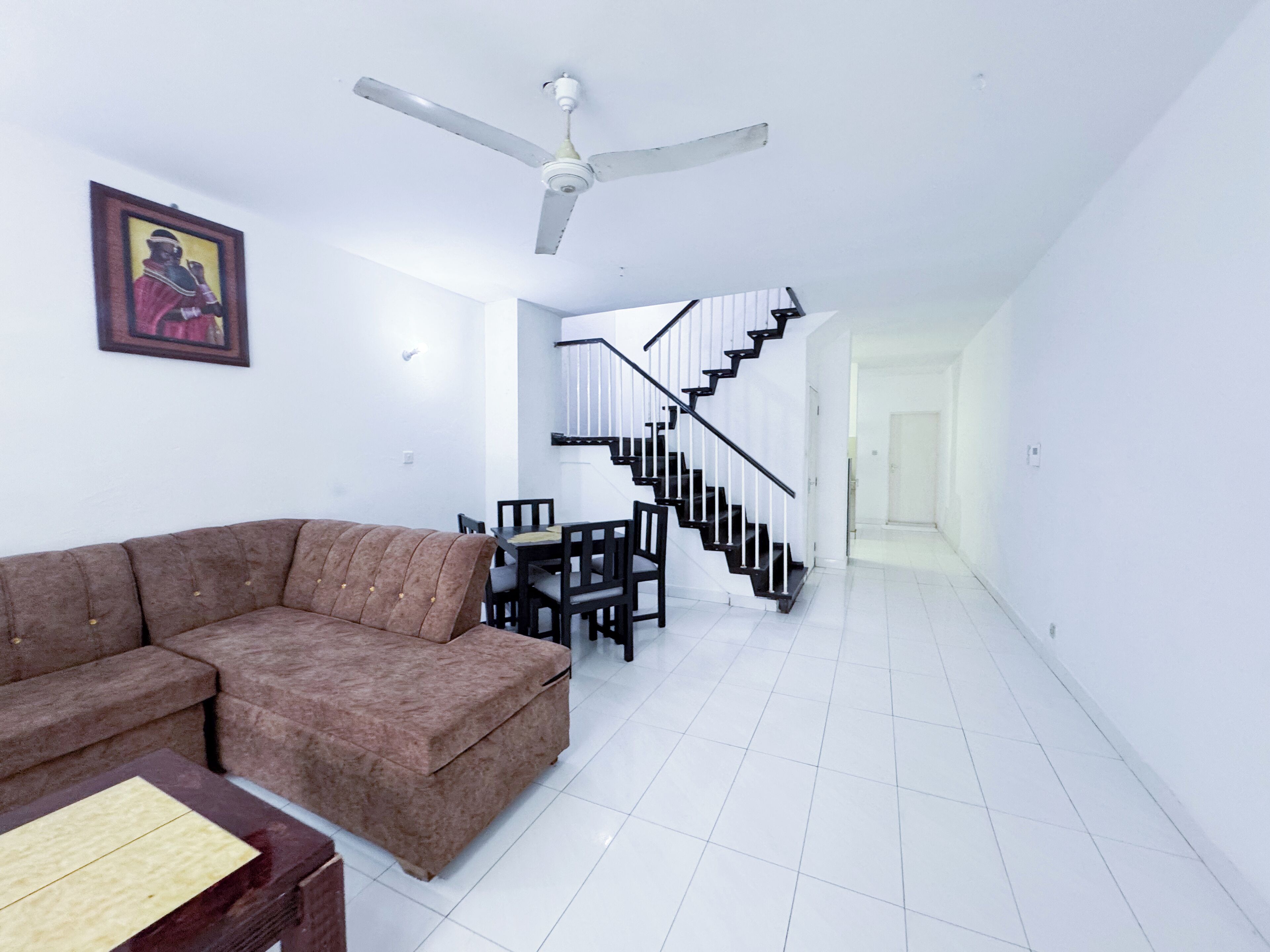 A living room with a brown sofa, a dining table with chairs, a ceiling fan, and a staircase leading to another level.